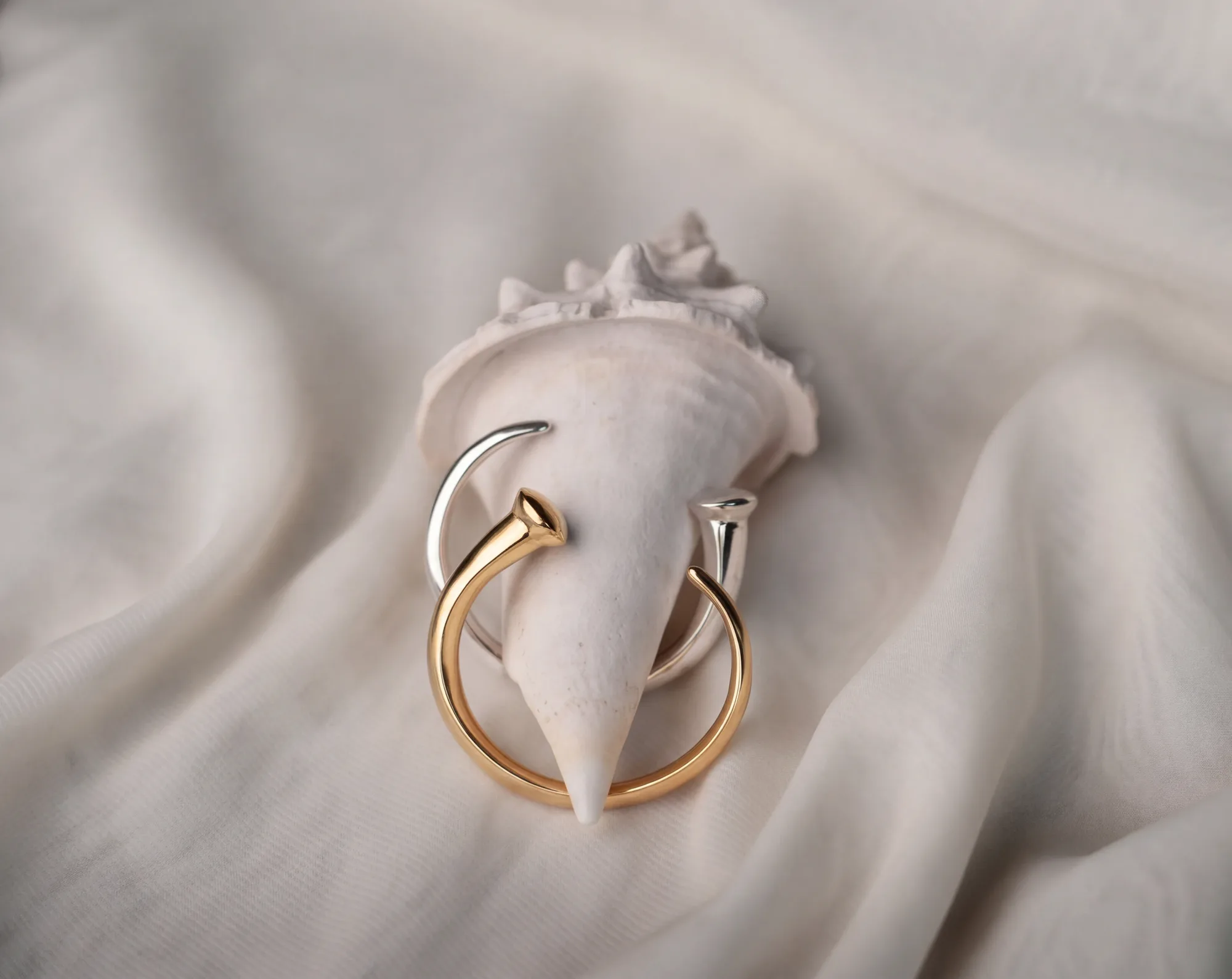 Deux bagues ouvertes, l’une en or et l’autre en argent, enroulées autour d’un coquillage blanc posé sur un drapé de tissu crème